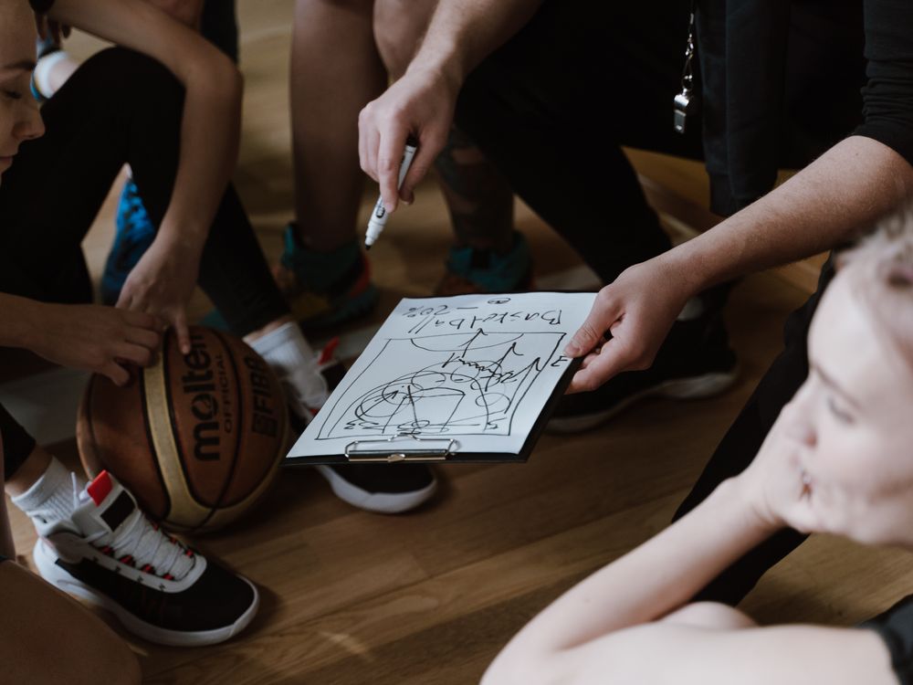 A coach sketching a play for a girls’ basketball team gathered on the court